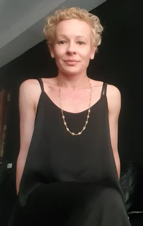 Una persona con cabello rubio corto y rizado, que viste un vestido negro sin mangas y un collar de oro, está sentada sobre un fondo oscuro.